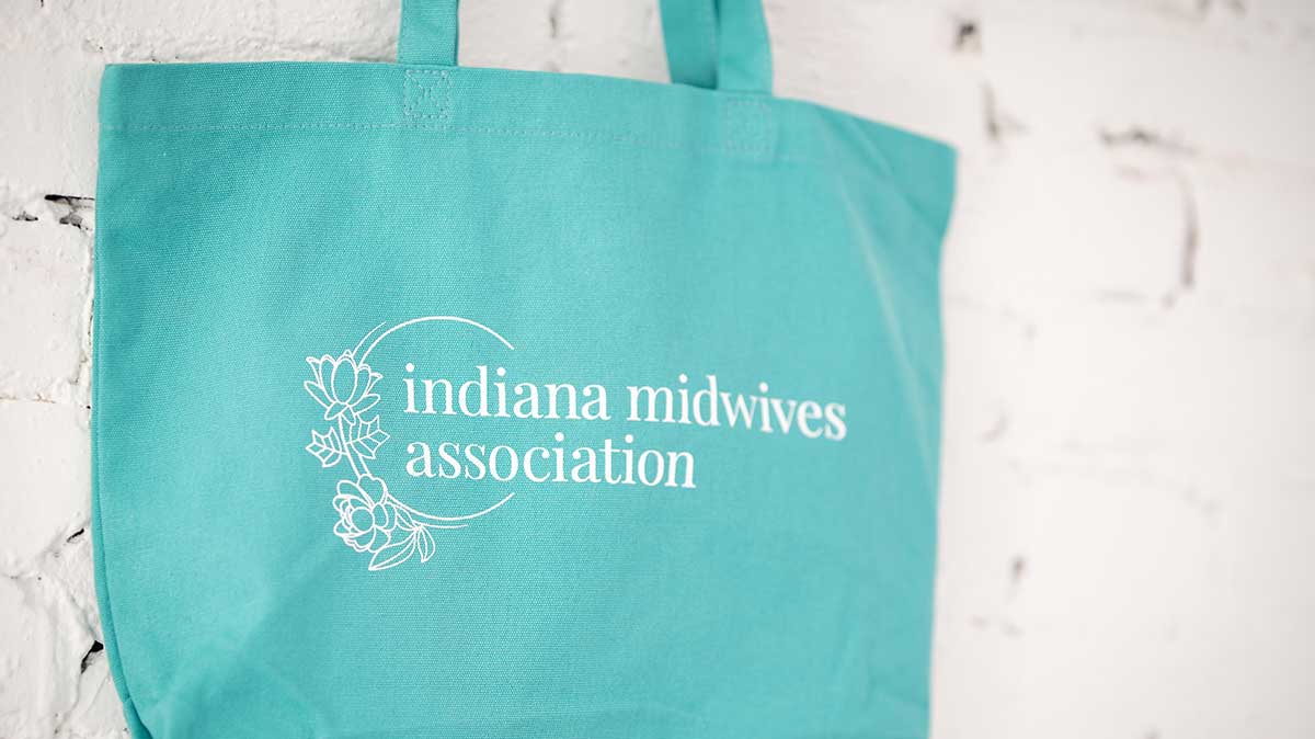 Blue tote bag hanging against a white brick wall - the logo is floral and says Indiana Midwives Association