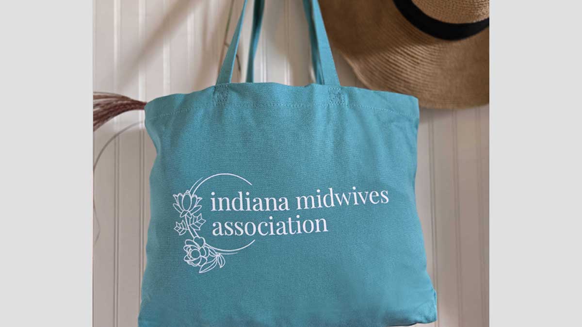 Blue tote bag with the white Indiana Midwives Association logo design. It's hanging in a white mudroom next to a hat.